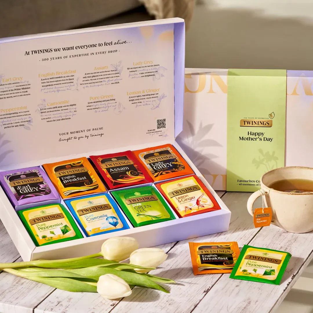 A box of assorted Twinings tea bags sits open, displaying various flavors. A cup of tea and tulips are nearby. The box is labeled “Happy Mother’s Day.”