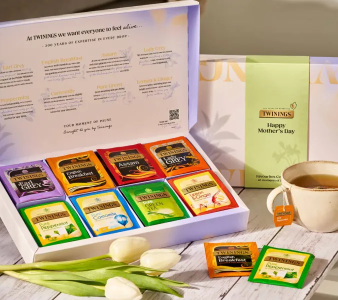 A box of assorted Twinings tea bags sits open, displaying various flavors. A cup of tea and tulips are nearby. The box is labeled “Happy Mother’s Day.”