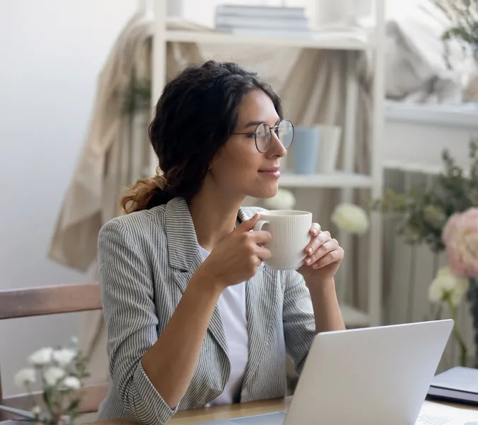 A woman, wearing glasses, sits at a desk in a home office, thoughtfully holding a mug. A laptop sits in front of her.