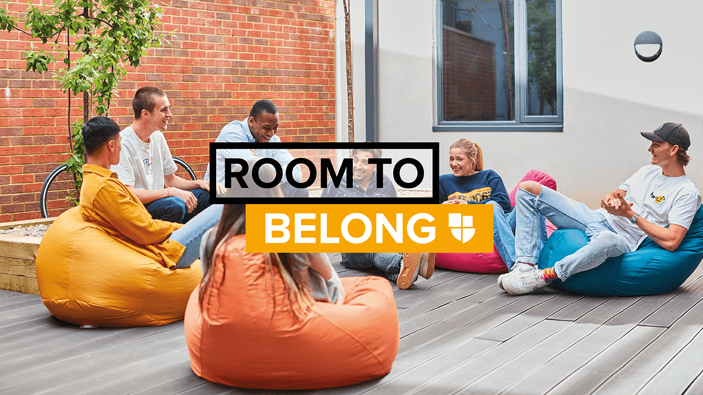 Young adults sit in beanbag chairs, socializing in an outdoor patio area. ROOM TO BELONG is overlaid on the image.