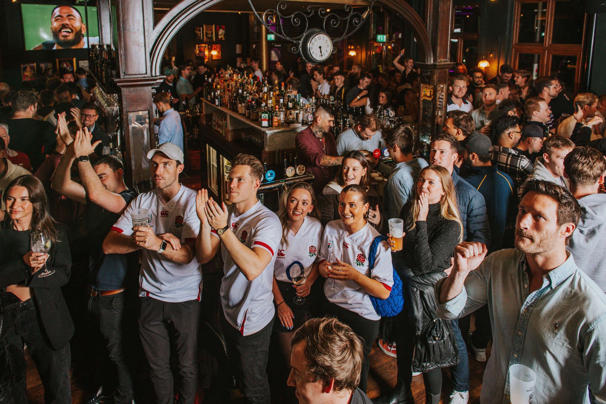 Rugby fans in white jerseys applaud, surrounded by a lively pub crowd at a bar.