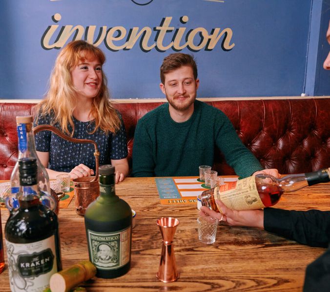 Two people sit on a red leather banquette, a bartender pours liquor. The wall behind them features the word "invention".