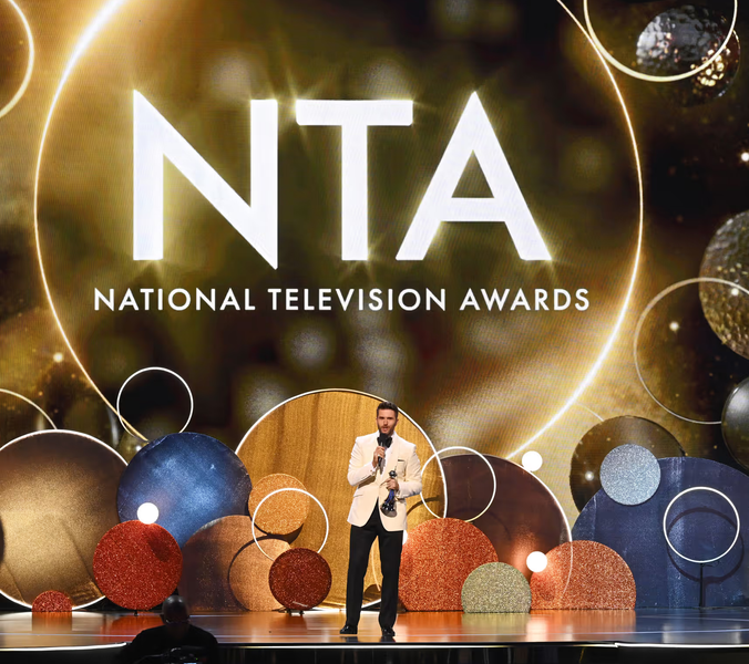 A man accepts an award on an awards show stage. The backdrop displays "NTA NATIONAL TELEVISION AWARDS."