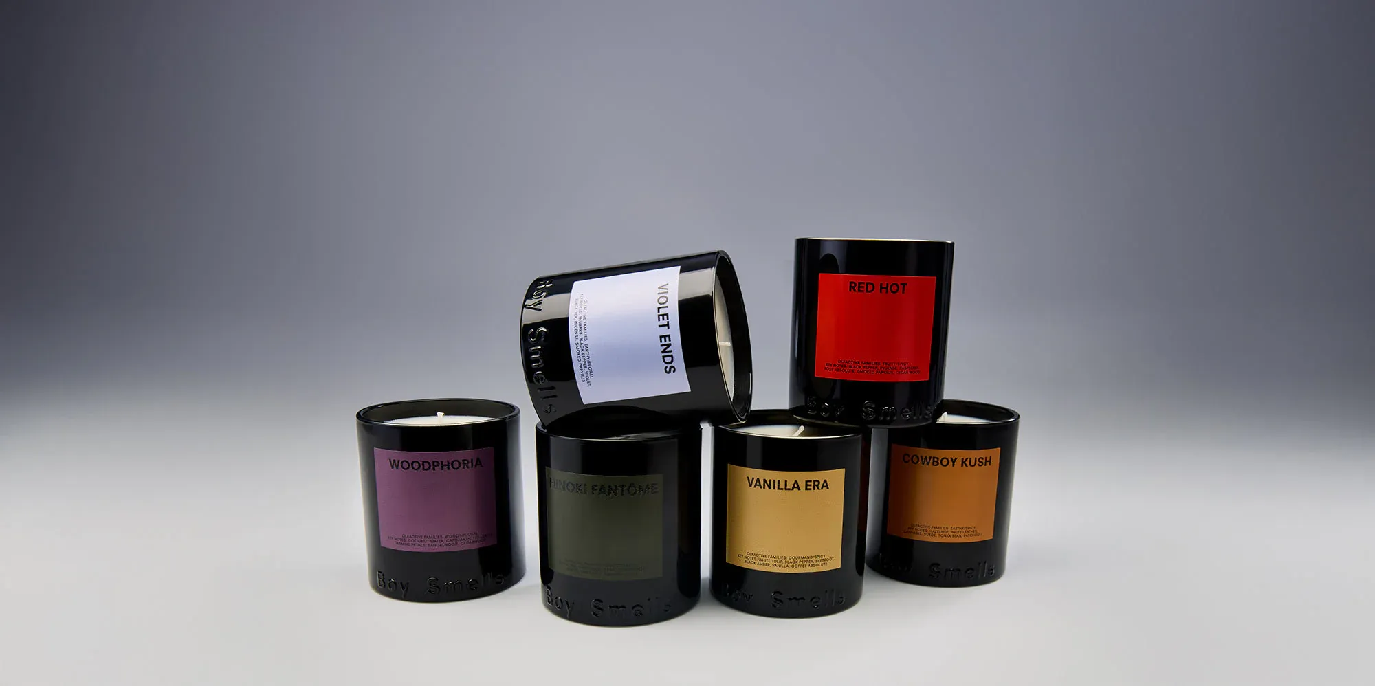 Five black Boy Smells candles, unlit, are stacked in a pyramid on a light surface against a gray backdrop. The labels indicate scents: Woodphoria, Violet Ends, Red Hot, Hinoki Fantôme, and Cowboy Kush. Vanilla Era is visible on the bottom candle.