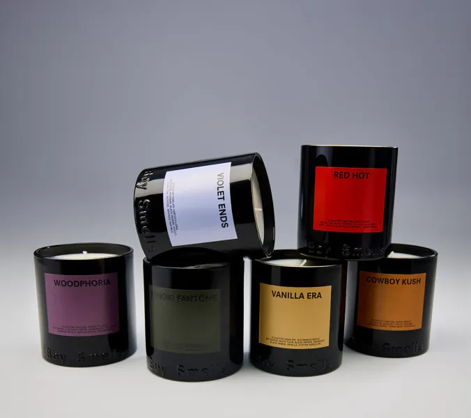 Five black Boy Smells candles, unlit, are stacked in a pyramid on a light surface against a gray backdrop. The labels indicate scents: Woodphoria, Violet Ends, Red Hot, Hinoki Fantôme, and Cowboy Kush. Vanilla Era is visible on the bottom candle.