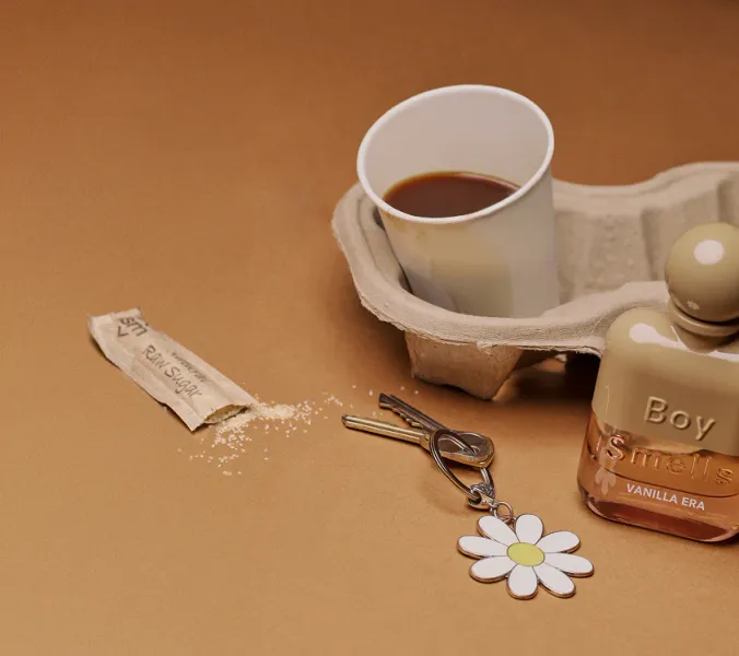 A Boy Smells Vanilla Era perfume and its travel-size counterpart rest on a cardboard coffee cup holder. A coffee cup, raw sugar packet, and keys with a daisy keychain are nearby on a brown surface.