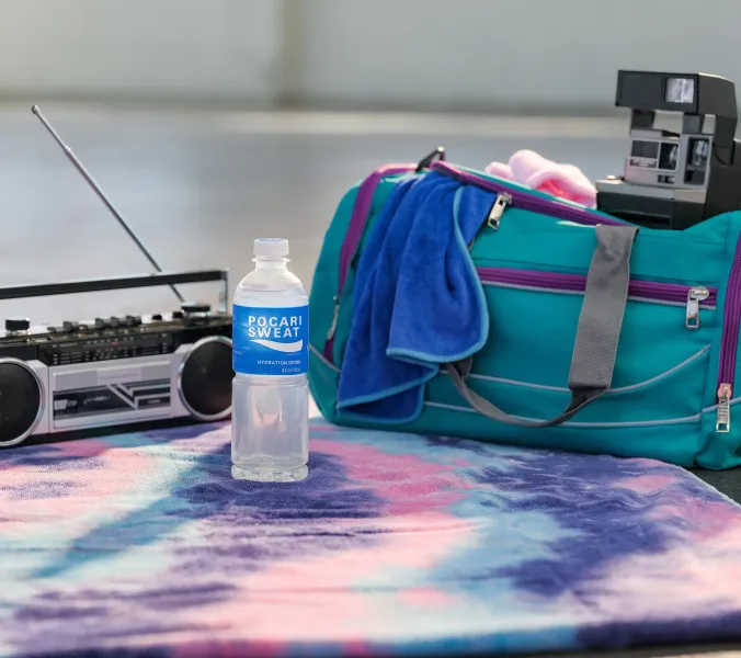 A Pocari Sweat hydration drink bottle rests on a tie-dye towel near a boombox, teal gym bag, and an instant camera.