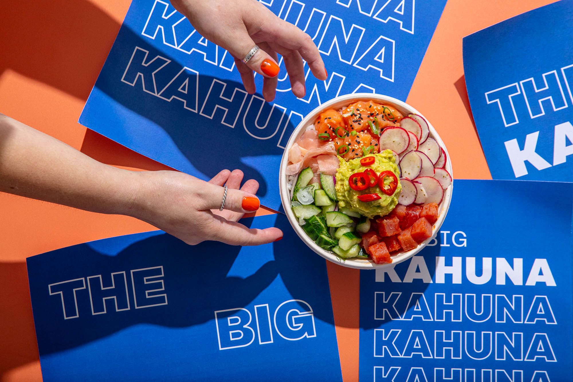 Hands reach for a poke bowl filled with rice, salmon, avocado, and radish. It sits on blue placemats with the words "THE BIG KAHUNA" repeated.