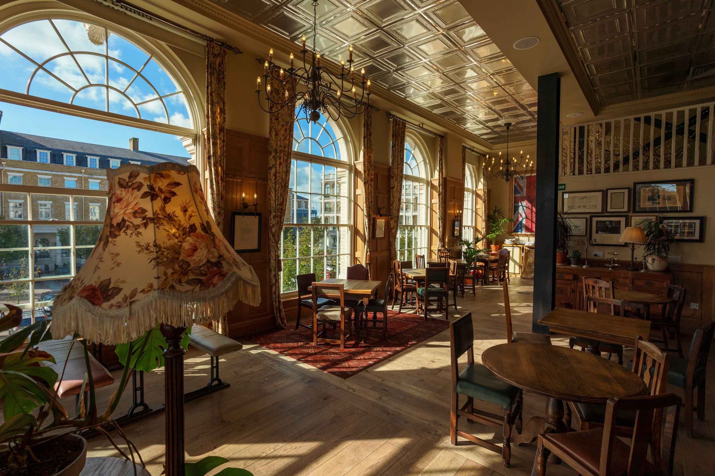 A floral lamp sits illuminating a sunlit, wood-floored pub. Tables and chairs are arranged throughout the room, which features large arched windows and a tin-tile ceiling. A Union Jack flag hangs on the wall.