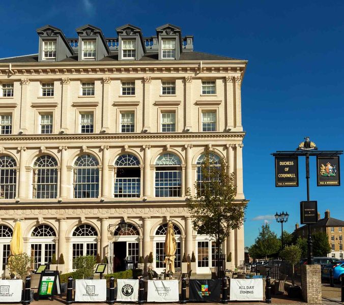 A large, tan building with many arched windows sits in a town square. The building is a pub called ""Duchess of Cornwall,"" with outdoor seating. Cars are parked nearby.