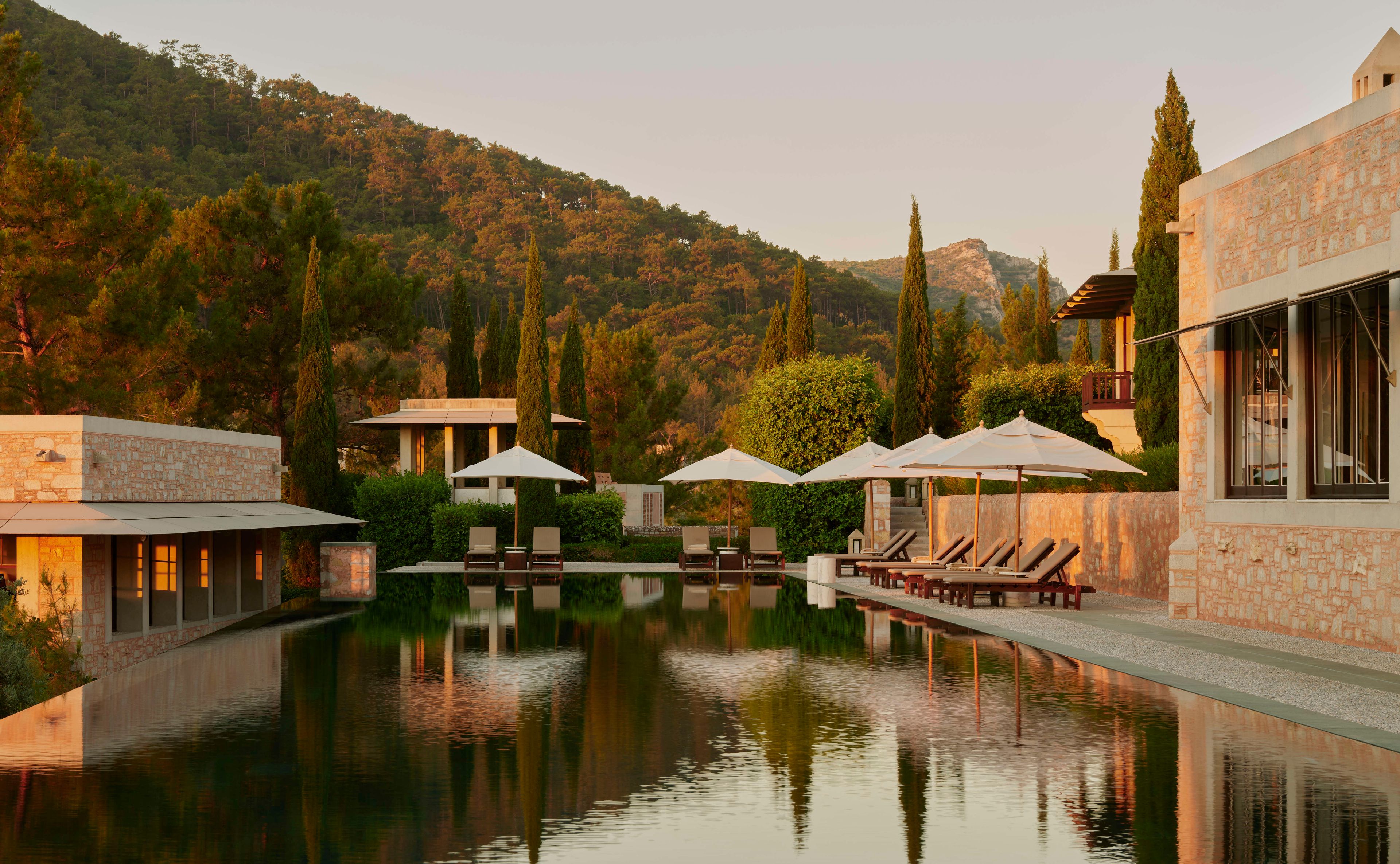 A reflecting pool sits beside a stone building, lined with lounge chairs and umbrellas. The scene is tranquil, set against a backdrop of a sunlit hillside covered in pine trees.