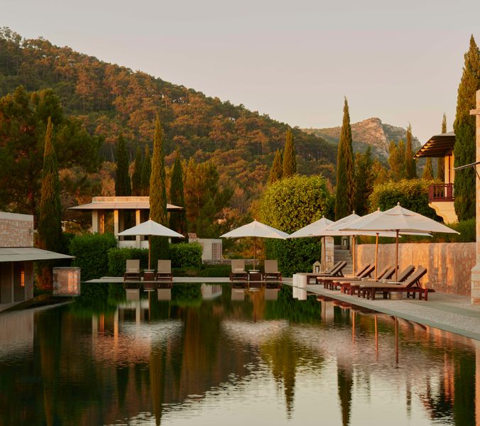A reflecting pool sits beside a stone building, lined with lounge chairs and umbrellas. The scene is tranquil, set against a backdrop of a sunlit hillside covered in pine trees.