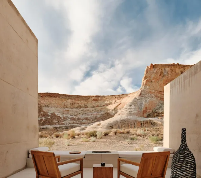 Two wooden chairs sit on a patio, facing a desert landscape with layered rock formations. A small wooden table is between them. A woven vase stands near a concrete wall.