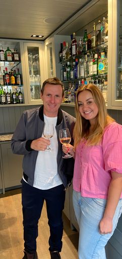 A man and woman stand, holding wine glasses, in a home bar with extensive liquor and glassware shelving.