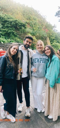 Four young adults stand together outdoors. Two women hold beverages. A man stands between them; one woman wears a “NEWPORT” sweatshirt. They are near a stone wall and greenery. "POV CAMERA 30 MAY 2024"