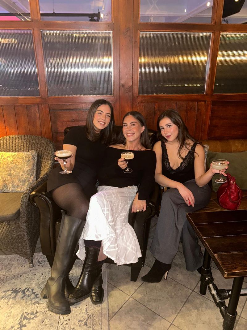 Three women sit together in armchairs, holding cocktails in a dimly lit bar.
