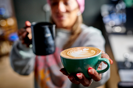 A barista presents a teal cup of latte art, holding a milk frothing pitcher, in a coffee shop.
