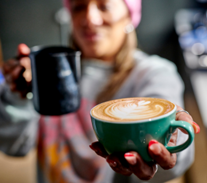 A barista presents a teal cup of latte art, holding a milk frothing pitcher, in a coffee shop.