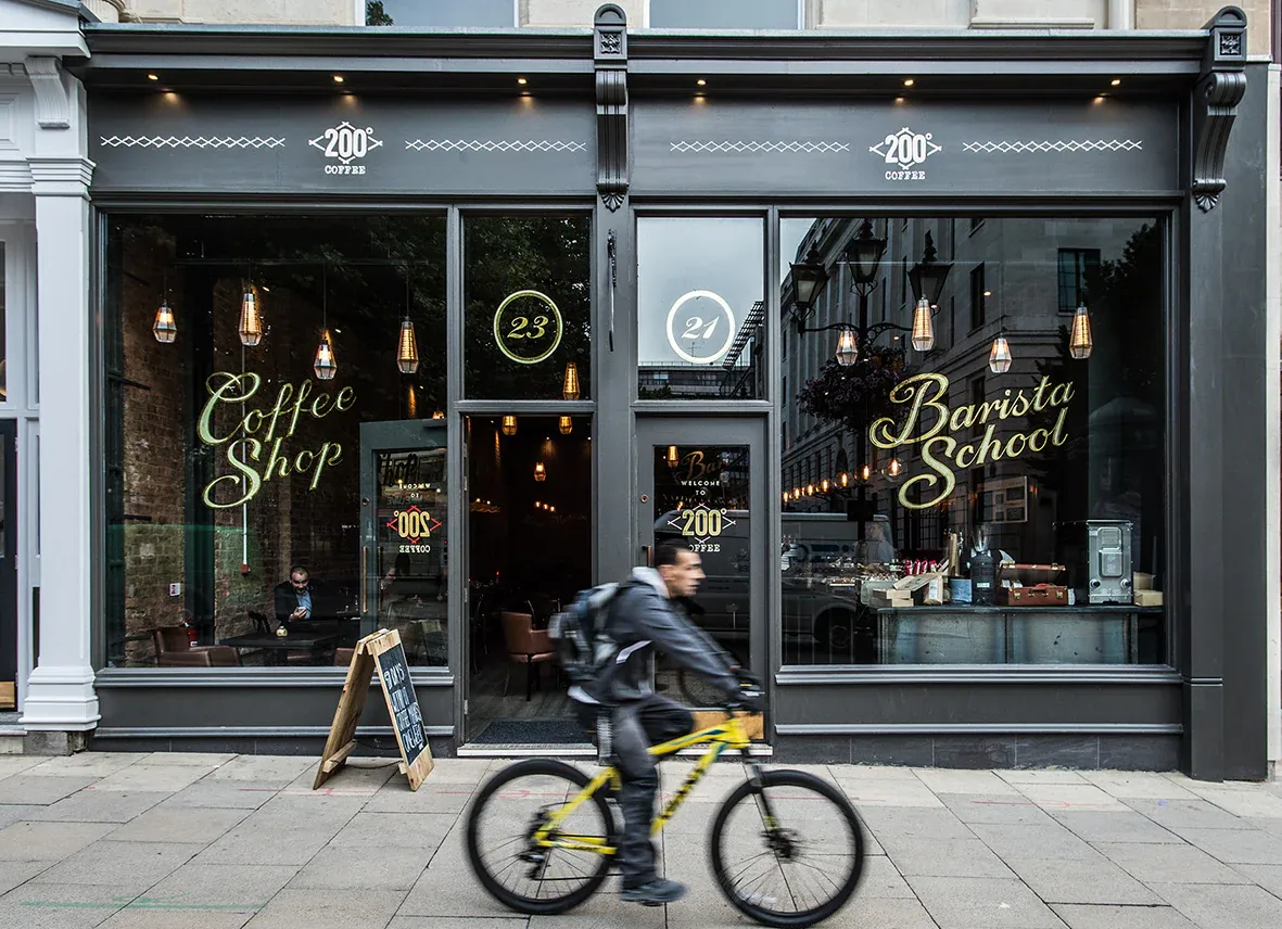 A cyclist rides past a dark-grey storefront. The shop's windows display ""Coffee Shop"" and ""Barista School"" signage. The address numbers 23 and 21 are visible. ""
