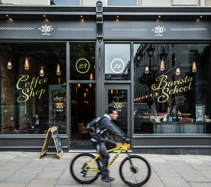 A cyclist rides past a dark-grey storefront. The shop's windows display ""Coffee Shop"" and ""Barista School"" signage. The address numbers 23 and 21 are visible. ""