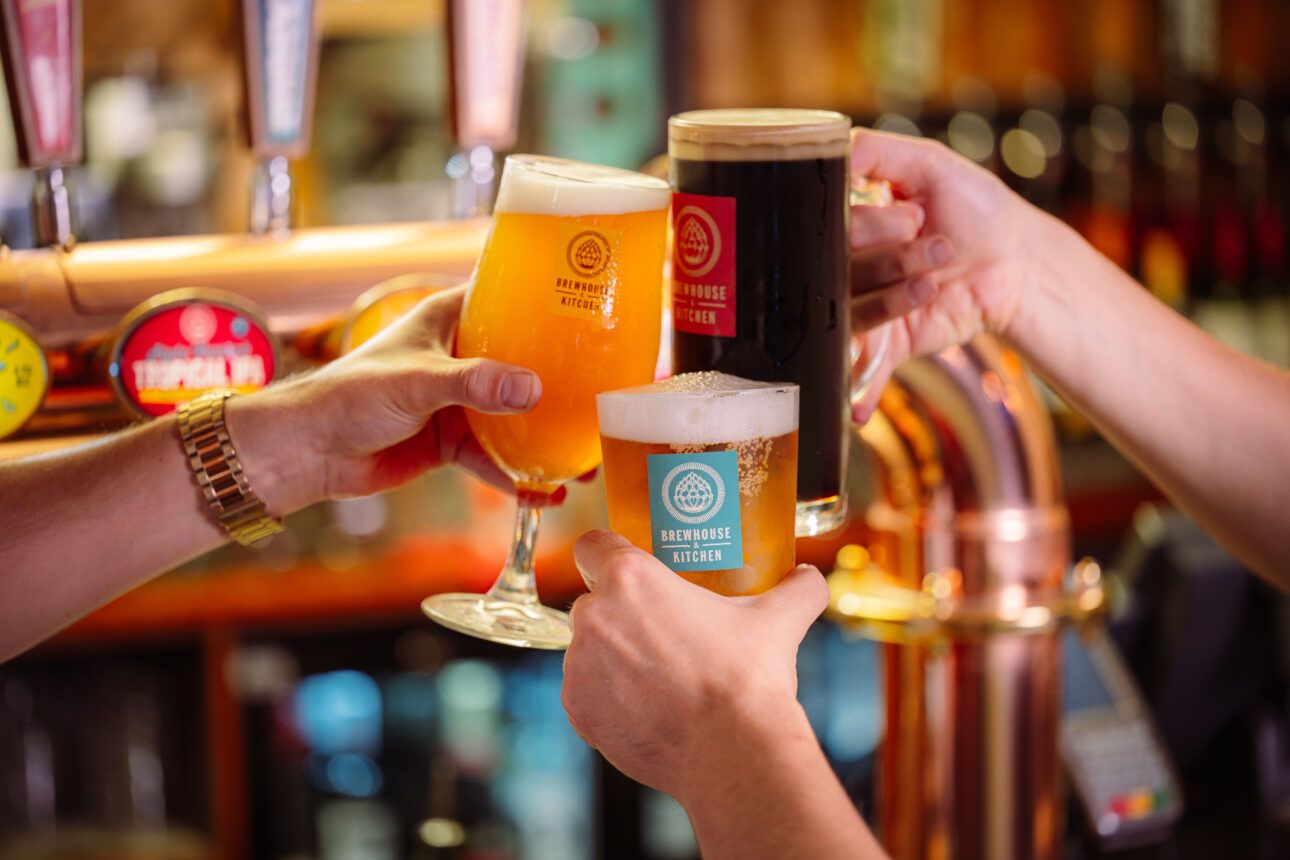 Three beer glasses—one pint, two stemmed—are raised in a toast at a bar. The glasses feature "BREWHOUSE KITCHEN" logos.