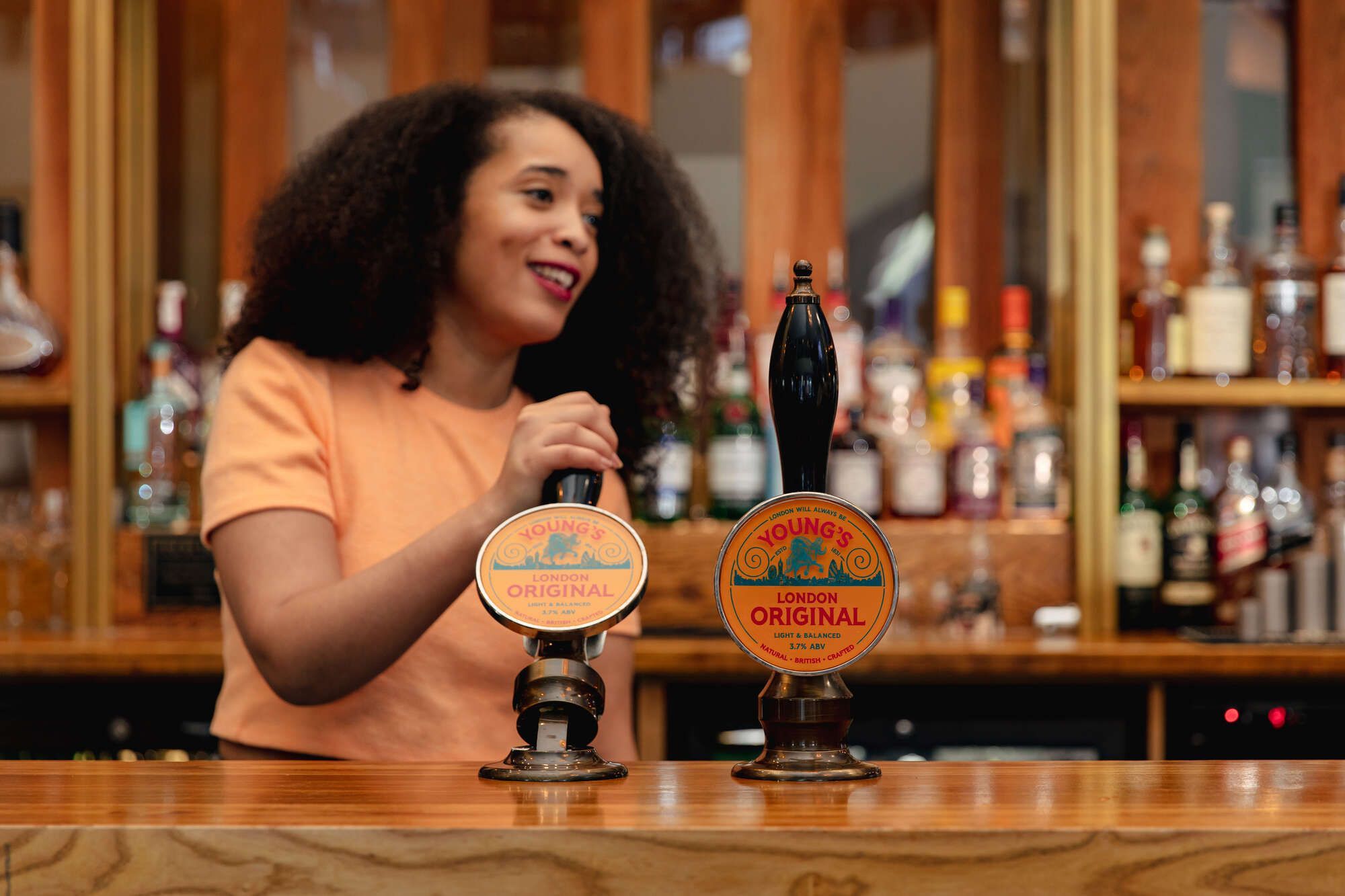 A bartender, partially visible, interacts with Young's London Original beer taps at a bar. The taps display "Young's London Original," "Light & Balanced," and "3.7% ABV."