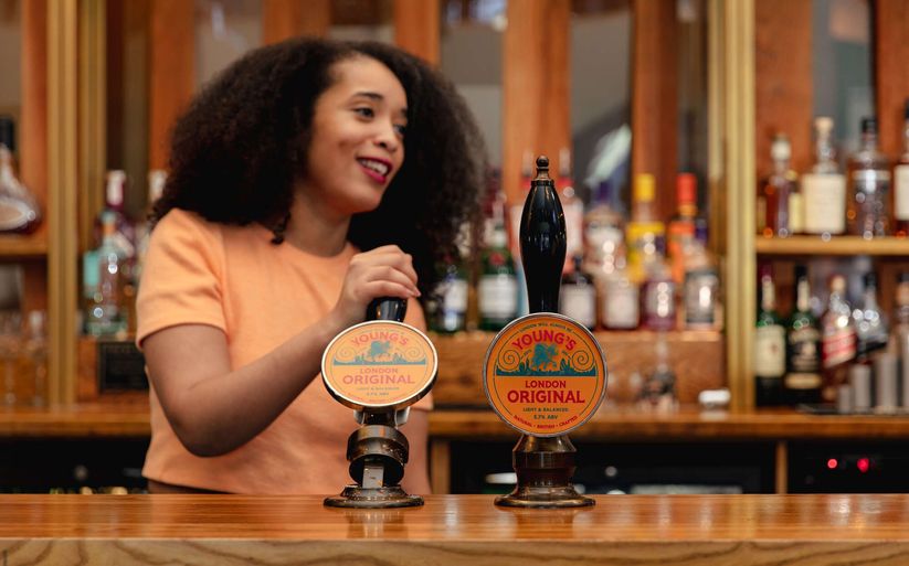 A bartender, partially visible, interacts with Young's London Original beer taps at a bar. The taps display "Young's London Original," "Light & Balanced," and "3.7% ABV."